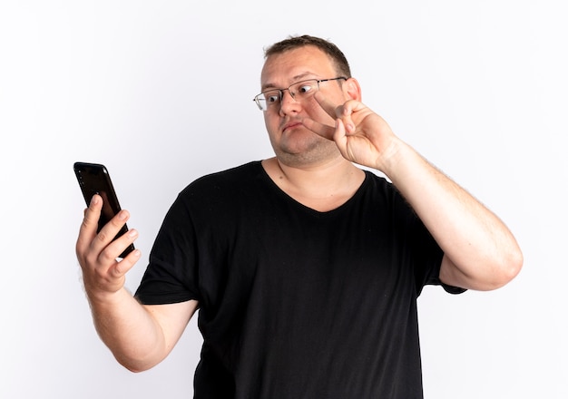 Hombre con sobrepeso en gafas con camiseta negra mirando la pantalla de su teléfono inteligente en línea mostrando el signo de la victoria de pie sobre la pared blanca