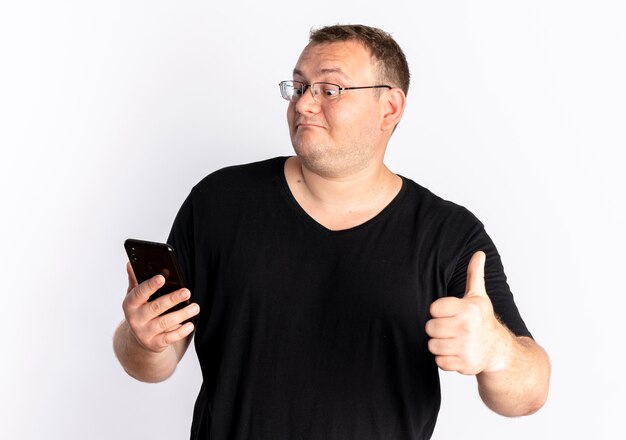 Hombre con sobrepeso en gafas con camiseta negra mirando la pantalla de su teléfono inteligente mostrando los pulgares para arriba de pie sobre la pared blanca