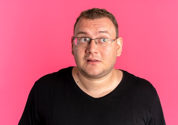 Hombre con sobrepeso en gafas con camiseta negra mirando sorprendido y feliz sobre rosa