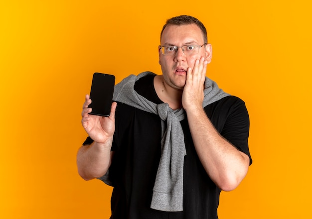 Hombre con sobrepeso en gafas con camiseta negra mostrando smartphone asombrado y sorprendido de pie sobre la pared naranja
