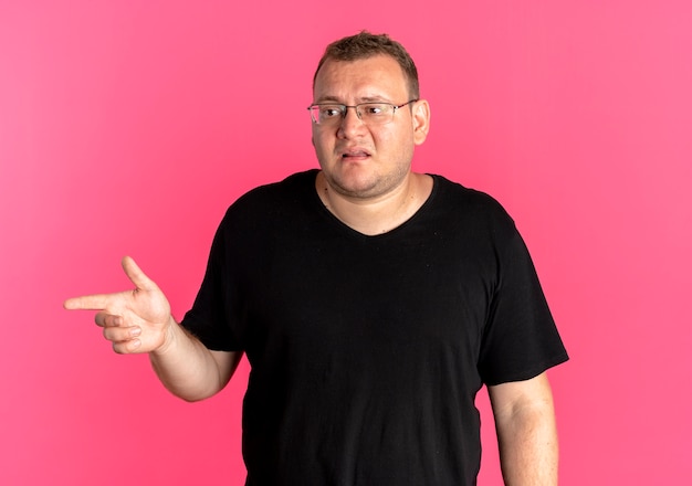 Hombre con sobrepeso en gafas con camiseta negra que parece confundido apuntando con el dedo índice hacia un lado sobre rosa