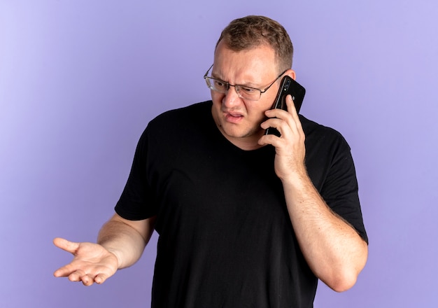 Hombre con sobrepeso en gafas con camiseta negra que parece confundido y disgustado mientras habla por teléfono móvil sobre azul
