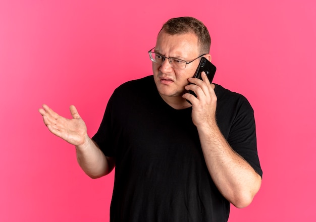 Hombre con sobrepeso en gafas con camiseta negra que parece confundido y disgustado mientras habla por teléfono móvil sobre rosa