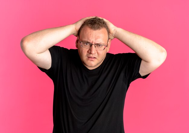 Hombre con sobrepeso en gafas con camiseta negra que parece confundido sosteniendo su cabeza con las manos sobre rosa