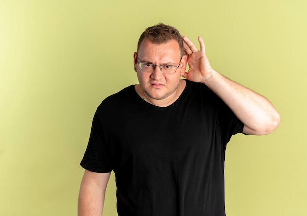 Hombre con sobrepeso en gafas con camiseta negra que parece confundido sosteniendo su palma cerca de la oreja tratando de escuchar de pie sobre una pared de luz
