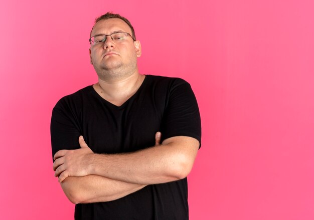 Hombre con sobrepeso en gafas con camiseta negra con rostro seroso con los brazos cruzados sobre el pecho de pie sobre la pared rosa