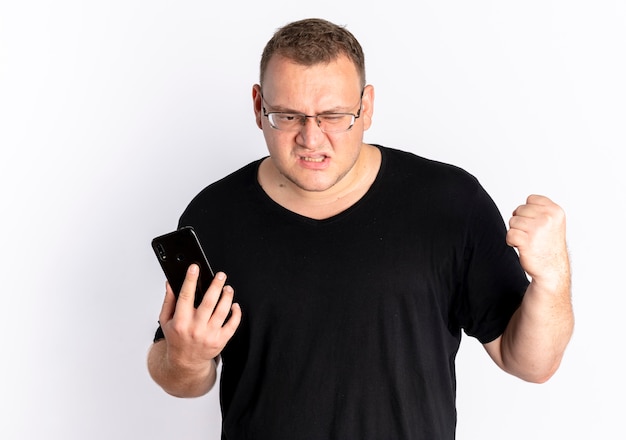 Hombre con sobrepeso en gafas con camiseta negra con smartphone apretando el puño con cara enojada de pie sobre la pared blanca