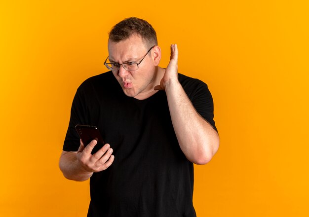 Hombre con sobrepeso en gafas con camiseta negra con smartphone apretando el puño gritando con expresión confusa de pie sobre la pared naranja