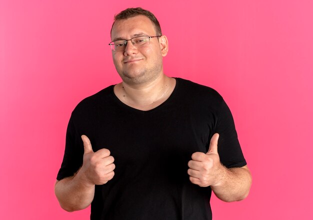 Hombre con sobrepeso en gafas con camiseta negra sonriendo mostrando los pulgares para arriba sobre rosa