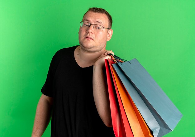 Hombre con sobrepeso en gafas con camiseta negra sosteniendo bolsas de papel mirando confundido sobre verde