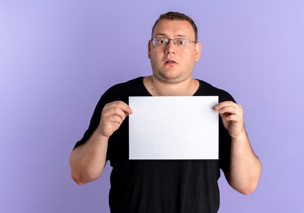 Hombre con sobrepeso en gafas con camiseta negra sosteniendo una hoja de papel en blanco lookign confundido de pie sobre la pared azul