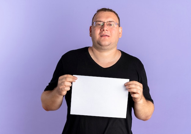 Hombre con sobrepeso en gafas con camiseta negra sosteniendo una hoja de papel en blanco con una sonrisa de pie sobre la pared azul
