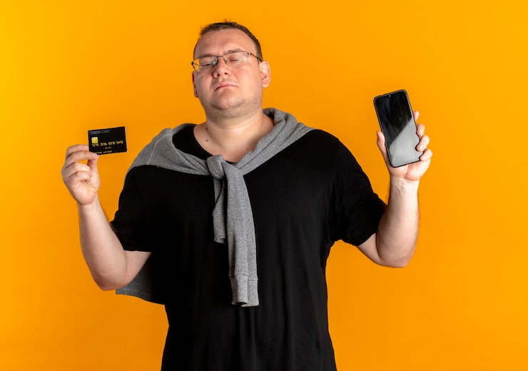 Foto gratuito hombre con sobrepeso en gafas con camiseta negra sosteniendo smartphone mostrando tarjeta de crédito mirando confiado sobre naranja