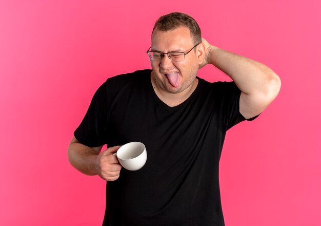 Hombre con sobrepeso en gafas con camiseta negra sosteniendo la taza de café divirtiéndose sacando la lengua rascándose la cabeza de pie sobre la pared rosa