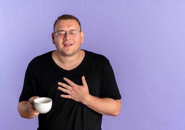 Hombre con sobrepeso en gafas con camiseta negra sosteniendo la taza de café sosteniendo la mano en el pecho sintiéndose agradecido de pie sobre la pared azul