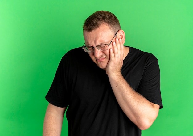 Foto gratuita hombre con sobrepeso en gafas vistiendo camiseta negra con aspecto enfermo tocando su mejilla con dolor de muelas sobre verde
