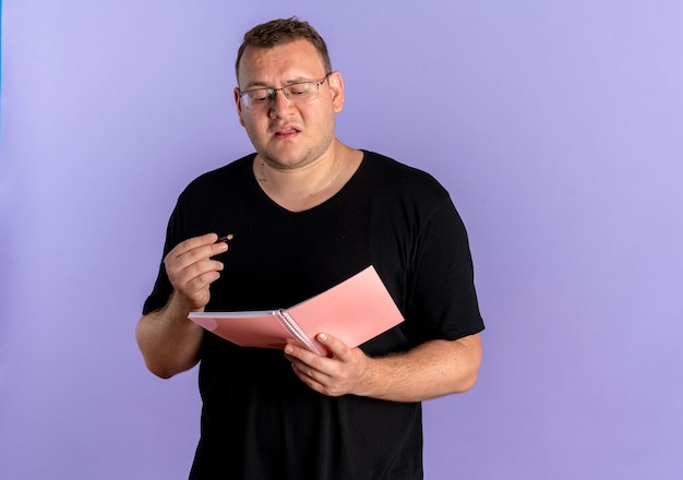 Hombre con sobrepeso en gafas vistiendo camiseta negra con cuaderno y bolígrafo mirando confundido de pie sobre la pared azul