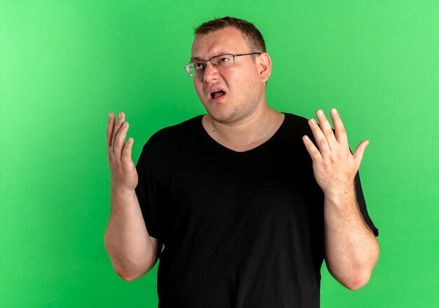 Hombre con sobrepeso en gafas vistiendo camiseta negra mirando disgustado gesticulando con las manos sobre verde