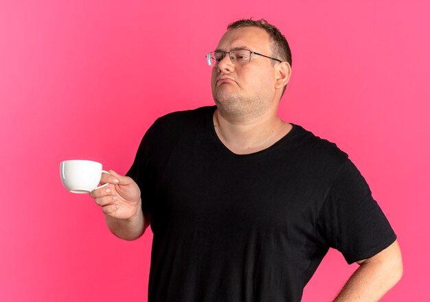 Hombre con sobrepeso en gafas vistiendo camiseta negra sosteniendo una taza de café mirando satisfecho sobre rosa