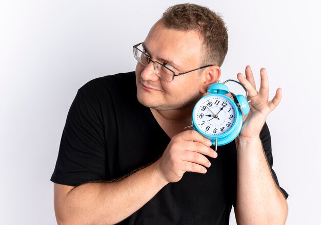 Hombre con sobrepeso vestido con camiseta negra con gafas sosteniendo un reloj despertador cerca de su oído tratando de escuchar el sonido de pie sobre la pared blanca