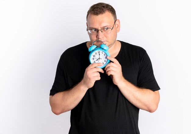 Hombre con sobrepeso vestido con camiseta negra con gafas sosteniendo reloj despertador mirando a un lado con expresión triste de pie sobre la pared blanca