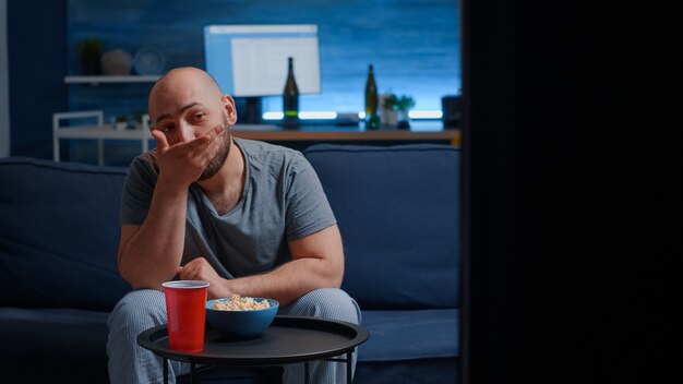 Hombre soltero disfrutando el fin de semana descansando delgado sentado en un cómodo sofá en la sala de estar solo comiendo palomitas de maíz ...