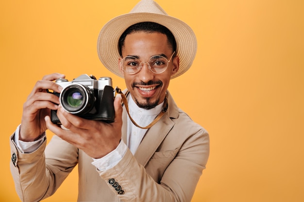 El hombre con sombrero beige está sosteniendo una cámara retro en la pared naranja