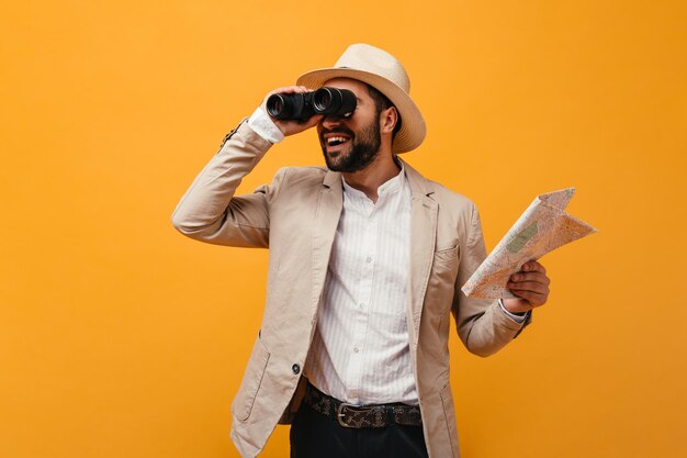 El hombre con sombrero mira hacia los binoculares y sostiene el mapa sobre fondo naranja