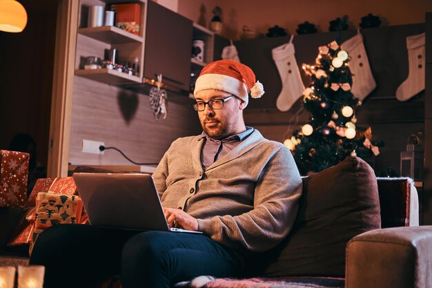 Hombre con sombrero de Papá Noel y anteojos vestidos con suéter cálido sentado en un sofá y trabajando en una laptop en Navidad.