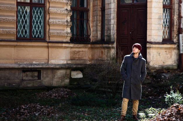 Hombre con sombrero rojo y abrigo gris está parado antes de la casa vieja