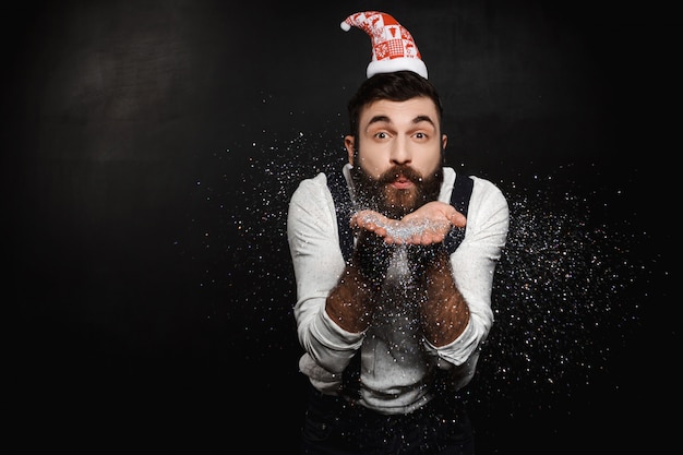 Hombre con sombrero de Santa Claus soplando brillo plateado sobre negro.