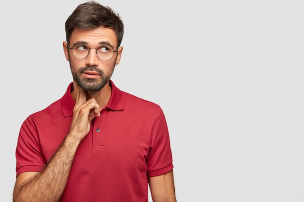 Hombre soñador pensativo con apariencia europea mira pensativamente hacia arriba, piensa en algo, analiza la situación de la vida, viste una camiseta roja, se para contra la pared blanca con espacio en blanco libre