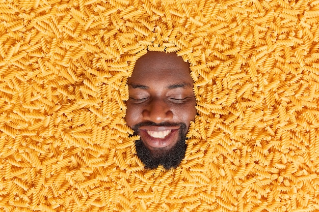 Foto gratuita el hombre sonríe ampliamente tiene dientes blancos perfectos cierra los ojos rodeado de pasta cruda preparada deliciosa comida de macarrones