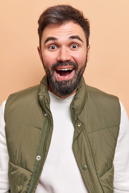 el hombre sonríe felizmente tiene expresión positiva se siente satisfecho oye excelentes noticias viste un jersey blanco con chaleco aislado en beige.