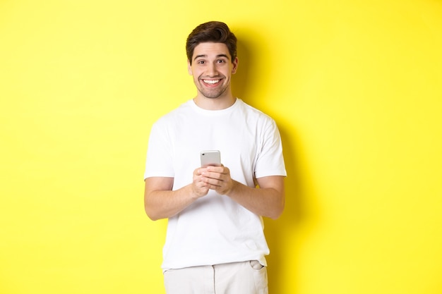 Hombre sonriendo y mirando feliz después de leer la oferta promocional en el teléfono inteligente, de pie contra el fondo amarillo en camiseta blanca.