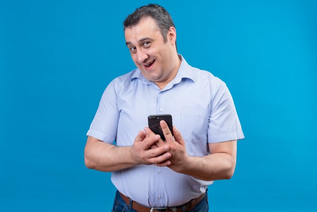 Hombre sonriente y alegre en camisa a rayas azul sosteniendo su teléfono móvil sobre un fondo azul.