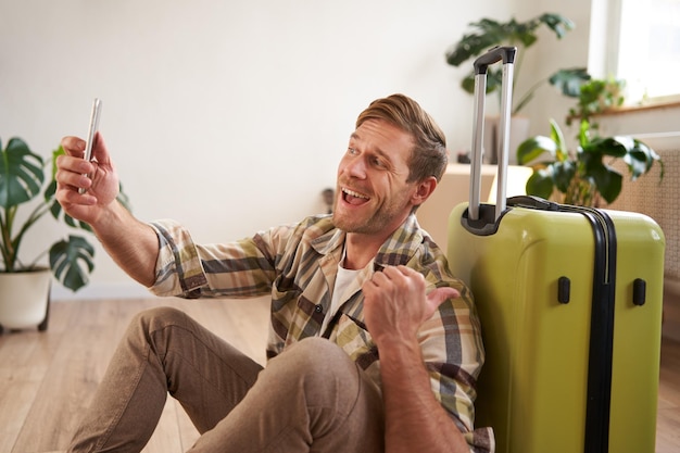 Foto gratuita un hombre sonriente y alegre con una maleta conversa por video usando su teléfono inteligente, se toma una selfie y se va de vacaciones.