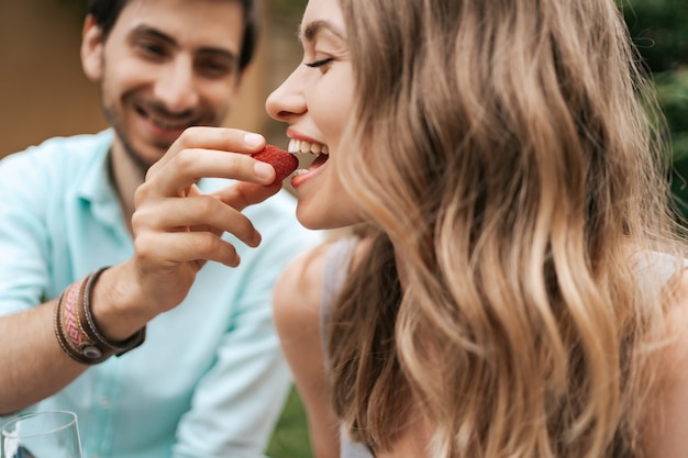 Hombre sonriente alimentando a su joven esposa feliz con fresa. Pareja alegre con hermosas sonrisas