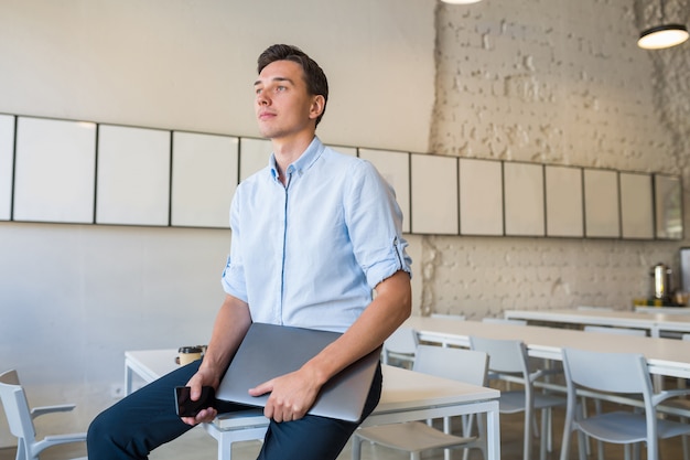 Hombre sonriente atractivo joven moderno sentado en la oficina abierta de coworking, sosteniendo el portátil