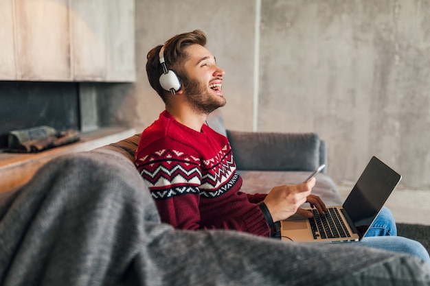 Hombre sonriente atractivo joven en el sofá en casa en invierno cantando música en auriculares, vistiendo suéter de punto rojo, trabajando en la computadora portátil, autónomo, emocional, riendo, feliz