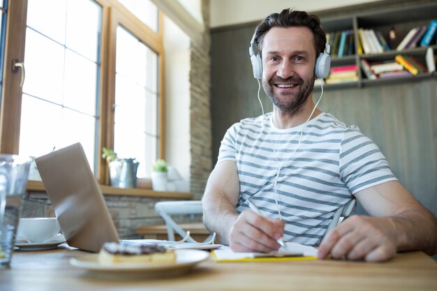 Hombre sonriente con auriculares escribiendo en su diario en la cafetería