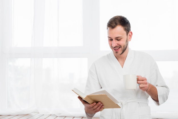 Hombre sonriente en bata de baño con libro y taza
