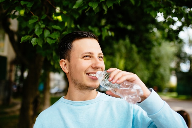 Hombre sonriente bebe agua de la botella en el fondo del árbol verde en día de verano