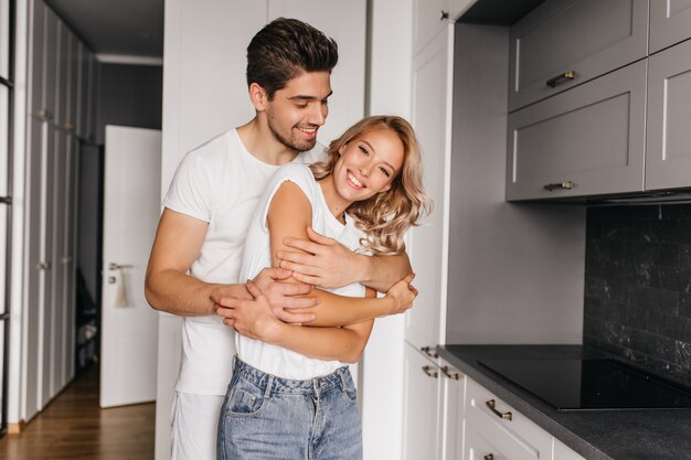 Hombre sonriente bronceado bailando con su esposa. Retrato interior de pareja abrazándose en acogedor apartamento.