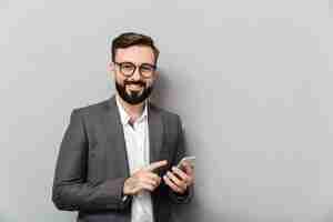 Foto gratuita hombre sonriente en camisa blanca escribiendo mensajes de texto o desplazamiento de alimentación en la red social con teléfono inteligente sobre gris