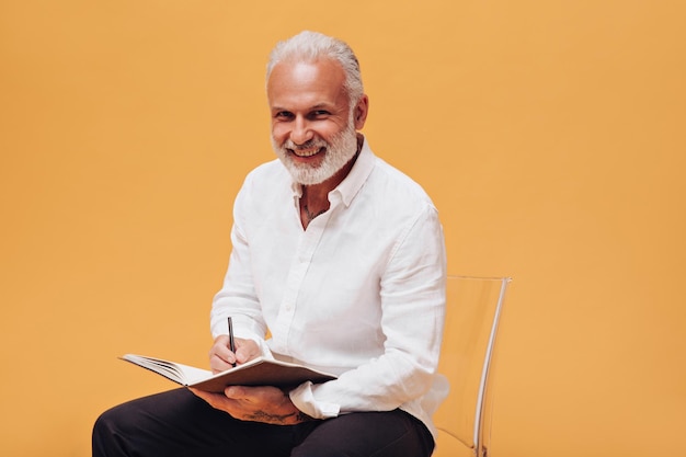 Hombre sonriente con camisa blanca mira a la cámara y sostiene un cuaderno Adulto canoso de buen humor se sienta en una silla y escribe en un bloc de notas