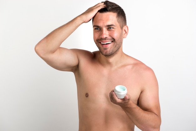 Hombre sonriente sin camisa que aplica cera en su cabello contra el fondo blanco