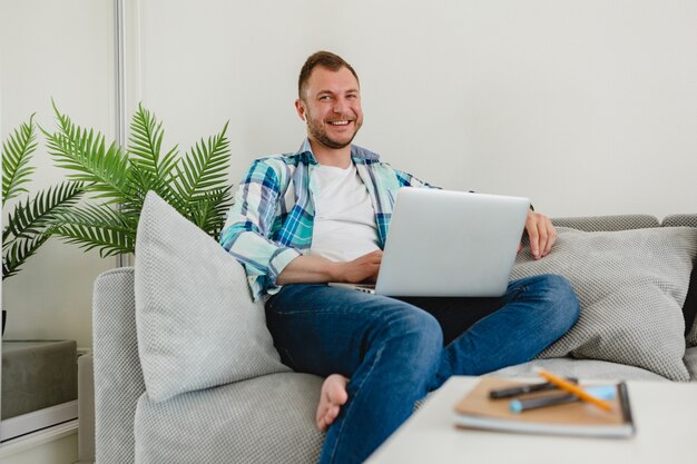 Hombre sonriente en camisa sentado relajado en el sofá en casa en la mesa trabajando en línea en la computadora portátil desde casa