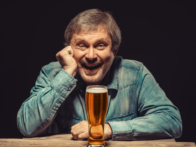 El hombre sonriente en camisa vaquera con vaso de cerveza