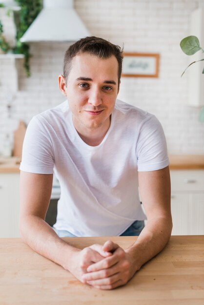 Hombre sonriente en la camiseta blanca que se sienta en la cocina